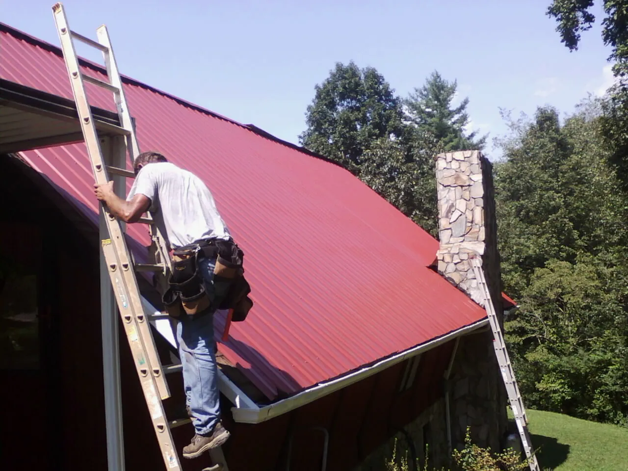 Roofer on a ladder beside a red metal roof and stone chimney — Soffit & Fascia Repair by Roofing Brothers in Carteret